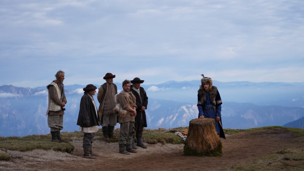 Grupa mężczyzn w tradycyjnych strojach góralskich podczas narady przy pniu drzewa na szczycie góry - kadr z filmu "Dziki",reż. Maciej Kawulski.